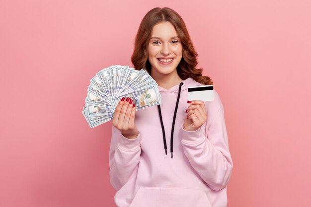 Retrato de una adolescente emocionada con el pelo rizado con capucha con tarjeta de crédito y mucho dinero, sorprendida por los depósitos en efectivo. Disparo de estudio interior aislado sobre fondo rosa