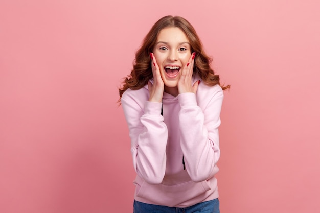 Retrato de una adolescente feliz sorprendida con capucha tocando la cara con las manos y sonriendo sinceramente con un estado de ánimo optimista Estudio interior aislado en un fondo rosa