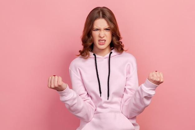 Retrato de una adolescente furiosa enfurecida con capucha de pie con los dientes apretados y las manos levantadas expresando una ira salvaje sintiéndose loca Foto de estudio interior aislada en un fondo rosa