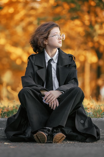 Retrato de una adolescente con gafas en impermeable negro sentada en una carretera asfaltada en el bordillo en el fondo del bosque otoñal