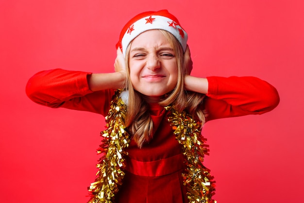 Retrato de una adolescente con un gorro de Papá Noel y oropel en el cuello cubriendo sus orejas con sus manos