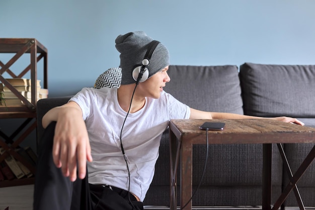 Retrato de un adolescente hipster con sombrero y auriculares sentados en el suelo en casa. Chico guapo de moda mirando hacia un lado, copie el espacio. Juventud, estilo de vida y ocio
