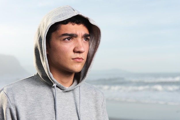 Retrato de un adolescente hispano guapo y serio que lleva una sudadera con capucha en la playa