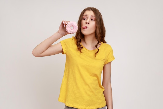 Retrato de una adolescente juguetona de cabello castaño con camiseta amarilla mirando donas y sacando la lengua divirtiéndose con comida dulce quiere lamer una toma de estudio interior aislada en un fondo gris