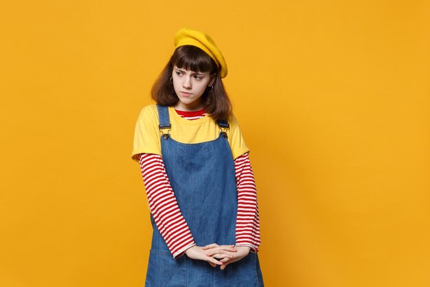 Retrato de una adolescente molesta preocupada con boina francesa, vestido de mezclilla mirando a un lado aislado en el fondo de la pared amarilla en el estudio. Emociones sinceras de la gente, concepto de estilo de vida. Simulacros de espacio de copia.