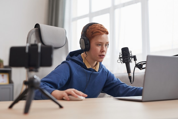 Retrato de adolescente pelirrojo jugando videojuegos con micrófono y cámara configurada para transmisión en línea, espacio de copia