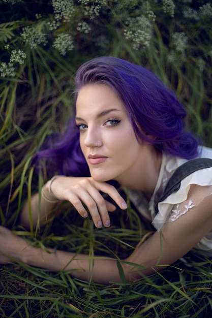 Retrato de una adolescente con el pelo morado y un pendiente en la nariz tumbado en la hierba en la naturaleza