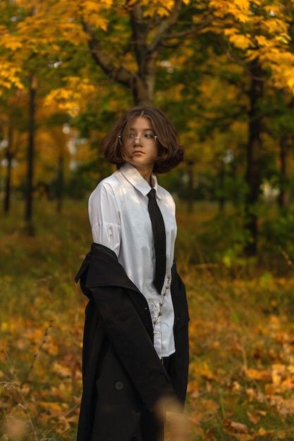 Retrato de una adolescente de pelo rizado con gafas y impermeable negro con corbata en el bosque de arce otoñal