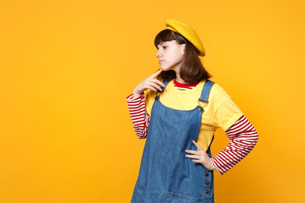 Retrato de una adolescente pensativa con boina francesa, vestido de mezclilla mirando a un lado, con la mano apoyada en la barbilla aislada en un fondo amarillo. Emociones sinceras de la gente, concepto de estilo de vida. Simulacros de espacio de copia.