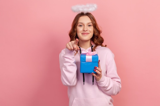 Retrato de una adolescente positiva de pelo rizado con capucha y halo en la cabeza sosteniendo una caja de regalo envuelta y señalando con el dedo a la cámara sonriendo sinceramente Foto de estudio interior aislada en fondo rosa