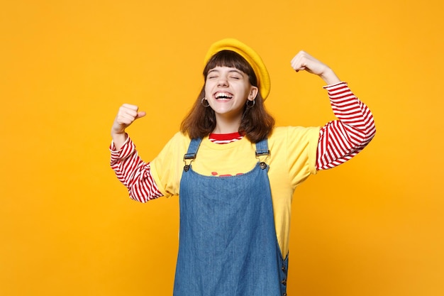 Retrato de una adolescente que se ríe con boina francesa, vestido de mezclilla que muestra bíceps, músculos aislados en el fondo de la pared amarilla en el estudio. Emociones sinceras de la gente, concepto de estilo de vida. Simulacros de espacio de copia.