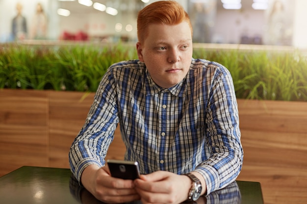 Un retrato de adolescente reflexivo con el pelo rojo y pecas vistiendo camisa a cuadros con teléfono móvil sentado en la mesa de madera