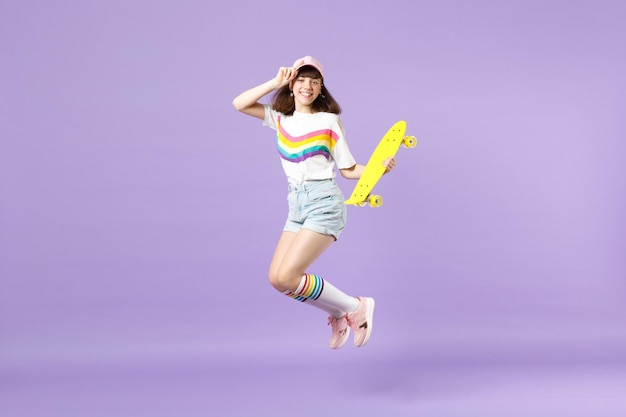 Retrato de una adolescente sonriente con ropa vívida sosteniendo una patineta amarilla, divirtiéndose, saltando aislada en un fondo de pared pastel violeta. Emociones sinceras de la gente, concepto de estilo de vida. Simulacros de espacio de copia.