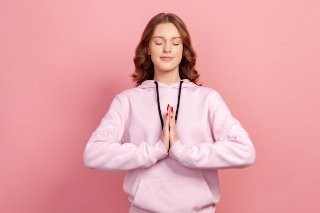 Retrato de una adolescente tranquila con capucha concentrando su mente manteniendo las manos gesto de namaste meditando la técnica de respiración del ejercicio de yoga reduce el estrés Toma de estudio interior aislada en fondo rosa