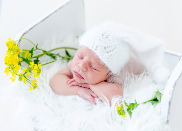 Retrato de adorable bebé recién nacido durmiendo boca abajo y sosteniendo sus manos debajo de las mejillas en la cama de madera blanca con flores amarillas. Lindo niño infantil con gorro de punto durmiendo