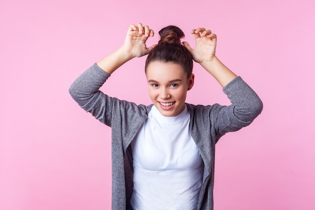 Retrato de una adorable y graciosa jovencita morena con peinado de moño en ropa informal que muestra orejas de conejo riéndose y divirtiéndose con un estado de ánimo infantil despreocupado en un estudio interior aislado en un fondo rosa