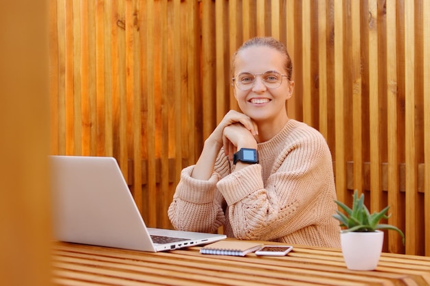 Retrato de una adorable mujer caucásica feliz con suéter beige descansando mientras trabaja en una laptop mirando la cámara con expresión positiva en la cámara posando en la oficina contra la pared de madera