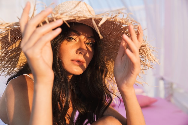 Retrato de adorable mujer morena en traje de baño y sombrero de paja sentado en la cama de la playa lounge en la mañana en el resort de verano