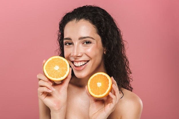 Retrato de adorable mujer semidesnuda con cabello largo sonriendo y sosteniendo trozos de naranja, aislado sobre fondo rosa