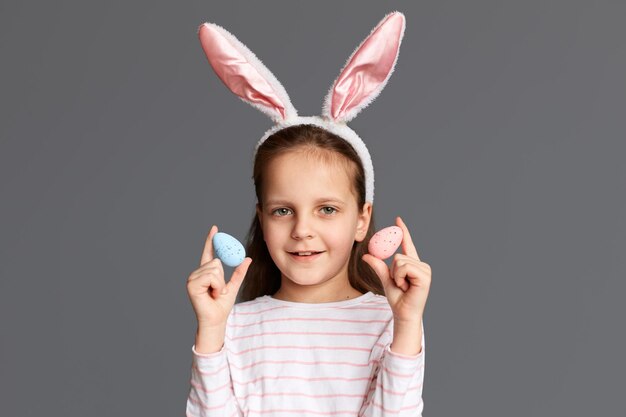 Retrato de una adorable niña caucásica con orejas de conejo sosteniendo dos huevos de Pascua mostrando su obra de arte de pintar testículos mirando la cámara aislada en un fondo gris