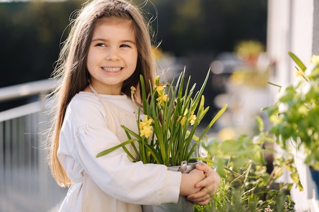 Retrato de una adorable niña sosteniendo narcisos en un balde de metal cerca de un macizo de flores Chica mira a la cámara y sonríe