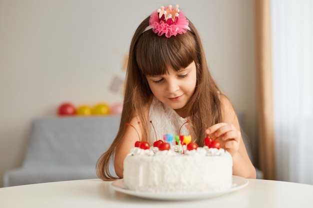 Retrato de una adorable niñita decorando pastel de cumpleaños para madre o padre, posando en una sala festiva con globos, mirando con concentraciones, poniendo cerezas encima del postre.