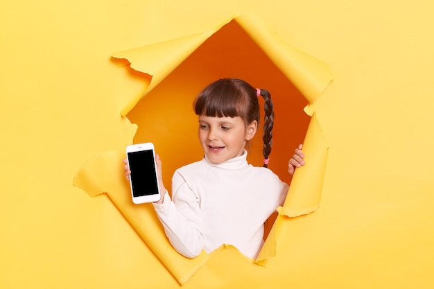 Retrato de una adorable niñita linda con trenzas con cuello de tortuga blanco posando en un agujero rasgado de una pared de papel amarillo que muestra un teléfono celular con una pantalla vacía con espacio de copia para publicidad