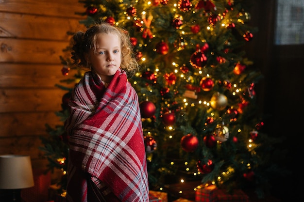 Retrato de una adorable niñita rubia y rizada envuelta en cuadros escoceses mirando a la cámara junto a un árbol de Navidad decorado con luces festivas brillantes