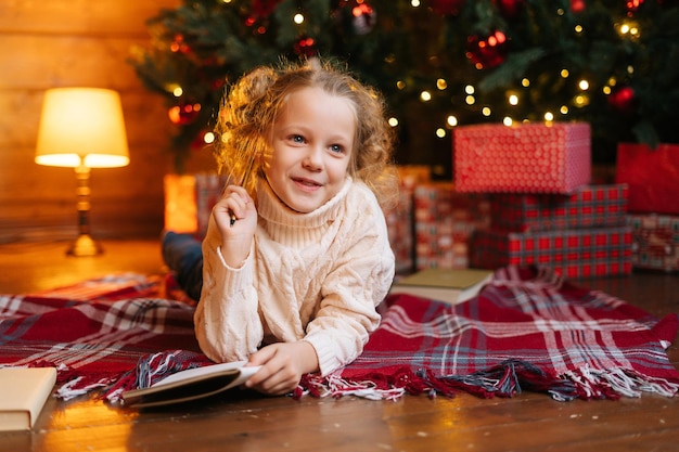 Retrato de una adorable niñita rubia soñando escribiendo una carta a Santa Claus tirada en el suelo sobre el fondo del árbol de Navidad y cajas de regalo