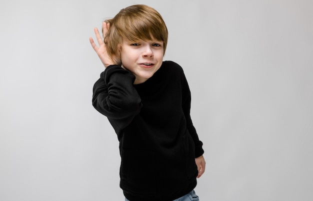 Retrato de adorable niño confiado de pie con las manos cerca de su cara escuchando en la pared gris