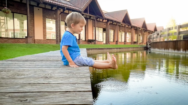 Retrato de adorable niño niño sonriente sentado en el río en la pequeña ciudad y sosteniendo los pies en el agua