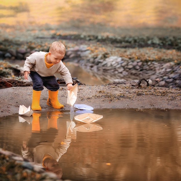 Retrato de un adorable niño pequeño con botas de goma lanza botes de papel en un charco en primavera Origami