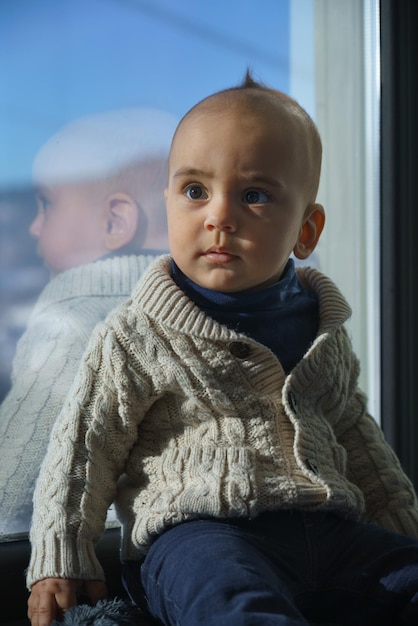 Foto retrato de un adorable niño con suéter beige sentado en el alféizar de la ventana