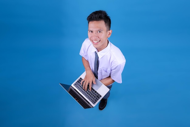 Retrato aéreo de un joven estudiante de secundaria indonesio usando una computadora portátil aislada sobre un fondo azul