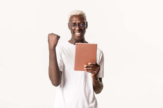 Retrato de afortunado chico afroamericano ganador, con gafas, sosteniendo una tableta digital y haciendo un puño en regocijo, sonriendo y triunfando, lograr el objetivo en la aplicación