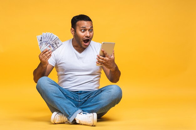 Retrato de afroamericano joven negro indio sentado en el suelo sosteniendo billetes de dólar aislados sobre fondo amarillo. Usando la computadora de la tableta.