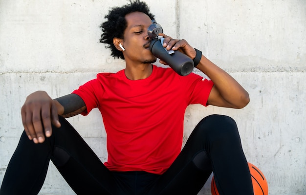 Retrato de agua potable de hombre afro atleta después de hacer ejercicio al aire libre. Deporte y estilo de vida saludable.