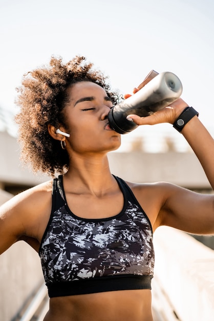 Retrato de agua potable de mujer afro atleta después de hacer ejercicio al aire libre. Deporte y estilo de vida saludable.