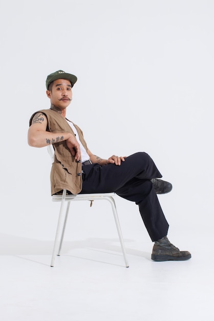 Retrato aislado de estudio de corte de cuerpo completo tiro asiático vintage de clase bigote cuello brazos tatuajes modelo de moda masculino en casual chaleco de safari de moda y gorra sentado en una silla blanca sobre fondo blanco