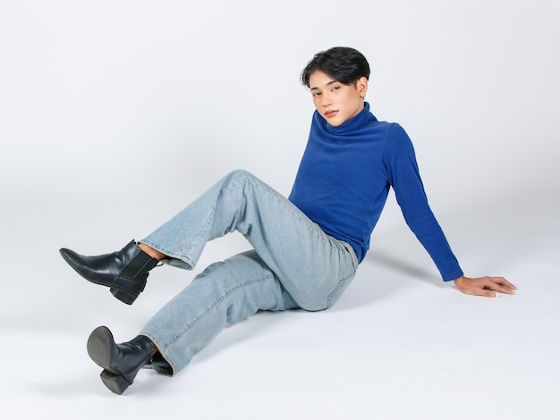 Retrato aislado de estudio de corte de todo el cuerpo asiático joven sexy delgado de moda LGBTQ modelo masculino gay en cuello alto camisa de manga larga jeans botas de cuero sentado tendido en el suelo posando sobre fondo blanco