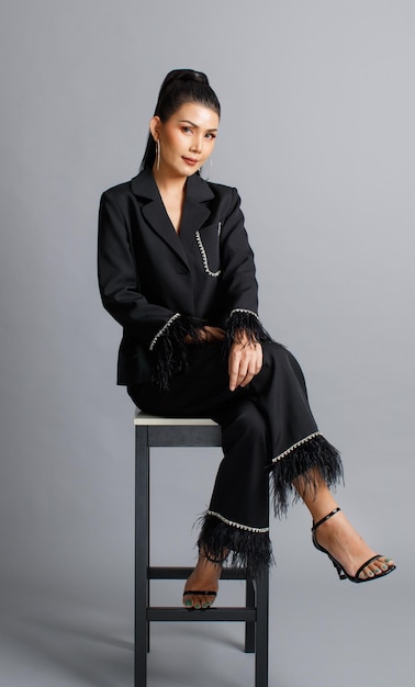 Retrato aislado en un estudio recortado de una elegante modelo de moda asiática con traje de piel de moda casual negro y tacones altos sentados con las piernas cruzadas en una silla mirando a la cámara posando en un fondo gris