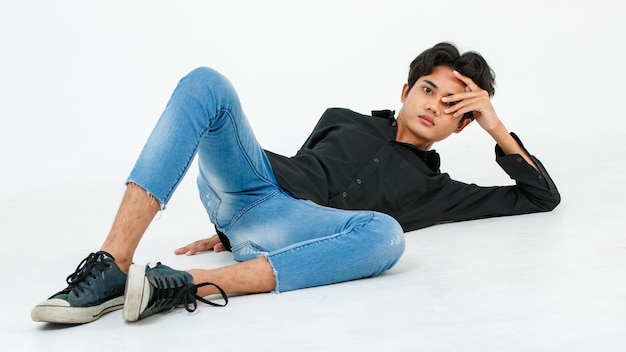 Retrato aislado de un estudio recortado de un joven asiático LGBTQ gay glamour modelo masculino con camisa negra casual y traje de jeans sentado estirando posando gesticulando en el suelo mirando a la cámara sobre fondo blanco