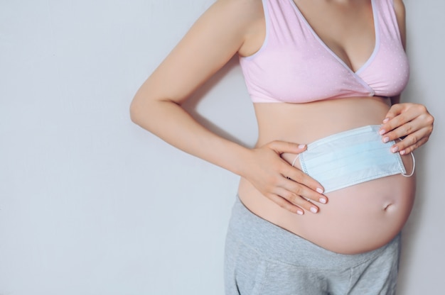 Retrato aislado de la mujer embarazada irreconocible que sostiene la máscara médica de la cara y que muestra a su bebé grande del vientre dentro. Concepto de embarazo, maternidad, nonato y expectativa. Gratis