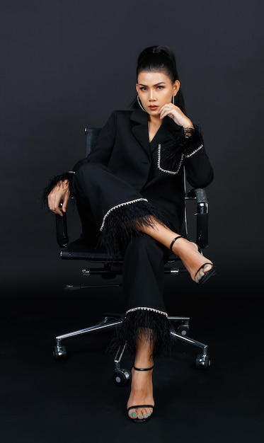 Retrato aislado recorte estudio tiro glamour femenino asiático elegante poderosa mujer de negocios de moda modelo en traje de moda casual negro y tacones altos sentado con las piernas cruzadas posando sobre fondo oscuro