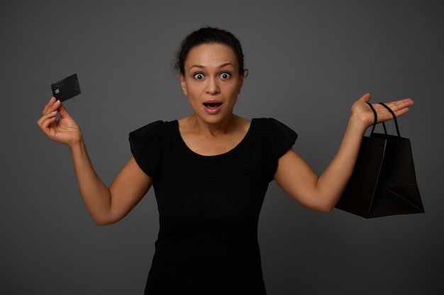 Retrato aislado sobre fondo gris con espacio de copia de una atractiva mujer asombrada vestida de negro y sosteniendo una tarjeta de crédito de descuento y una bolsa de compras, mirando a la cámara. Concepto de viernes negro