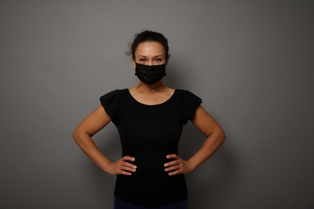 Retrato aislado sobre fondo gris de una mujer con máscara protectora médica negra mirando a la cámara con las manos en la cintura. Copie el espacio para publicidad. Concepto de viernes negro