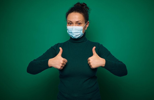 Retrato aislado sobre fondo verde de una mujer milenaria seria en máscara médica protectora, gesticulando, mostrando los pulgares hacia arriba mirando a la cámara. Copiar espacio publicitario