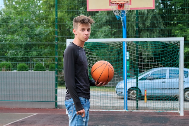 Retrato al aire libre de un adolescente con la pelota en la cancha de baloncesto de la calle, moda masculina de 16, 17 años mirando a la cámara. Estilo de vida activo y saludable, pasatiempos y ocio, concepto de adolescentes