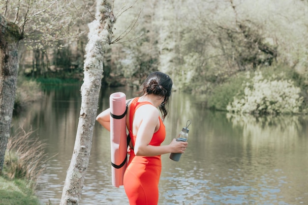 Retrato al aire libre de la afición del yoga de la mujer árabe que sonríe a la cámara al aire libre en la mañana en el parque cerca de un lago. Copie las imágenes del espacio, estirando y haciendo posiciones de yoga. Paños deportivos durante un día soleado.