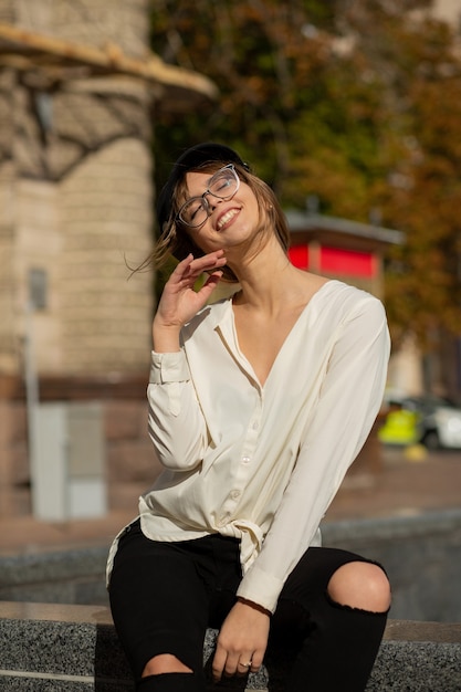 Retrato al aire libre de la alegre mujer morena lleva elegante gorra, gafas y blusa posando en la ciudad